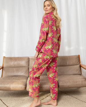 A woman smiles while sitting cross-legged on a bed, wearing the PRINTFRESH - BAGHEERA COTTON PAJAMA SET. The vibrant red and yellow hand screen-printed leopards complement the organic cotton pajamas, white bedding, brass lamp, and nightstand vase of white flowers.