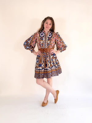 A woman poses against a plain background in the FARYLINA ANDI DRESS, a colorful bohemian style with long puff sleeves and a brown belt, styled with tan flats and long hair, one leg crossed in front of the other.