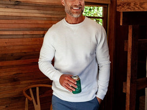 A smiling man with a short beard wears the CRIQUET - QUILTED CREW SWEATSHIRT IN OFF-WHITE by CRIQUET, holding a green can indoors near wood-paneled walls, a window, and a wooden chair.