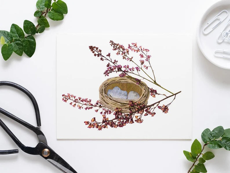 A small, woven green basket with light blue paper inside, surrounded by purple hydrangea sprigs on a white background. (Product: PETAL PEOPLE PRESS - NEST GREETING CARD by PETAL PEOPLE PRESS).