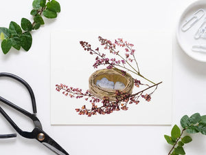 A small, woven green basket with light blue paper inside, surrounded by purple hydrangea sprigs on a white background. (Product: PETAL PEOPLE PRESS - NEST GREETING CARD by PETAL PEOPLE PRESS).