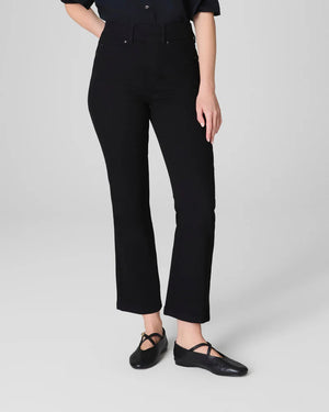 A woman stands against a light gray background, smiling with her hands behind her back. She wears the SPANX KICK FLARE JEAN IN CLASSIC BLACK, a high-waisted jean made from 4-way stretch denim, paired with a black button-up shirt and black flats.