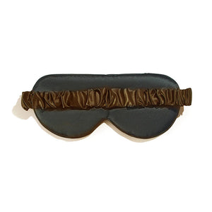 A woman with long, dark hair is lying on a bed adorned with a silky gold sheet. She is wearing a dark tank top and holding the BRANCHE - BELLE DE NUIT SILK EYE MASK in blue up to her forehead, smiling at the camera.