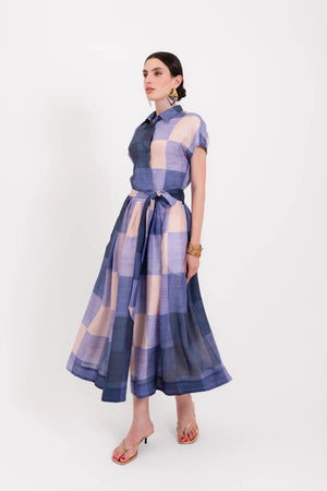 A woman wearing the LAVI - BELLELL Dress in Plaid by LAVI, a short-sleeve, collared shirt dress with a blue and beige plaid pattern and a tied waist, accessorized with gold earrings, bracelets, and open-toed heels, stands against a plain white background.