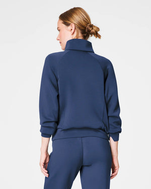A person with short hair is wearing a SPANX AIRESSENTIALS HALF ZIP sweater in navy blue, made from lightweight spacer fabric, paired with black pants. Their arms are crossed over their chest as they stand against a plain, light grey background.