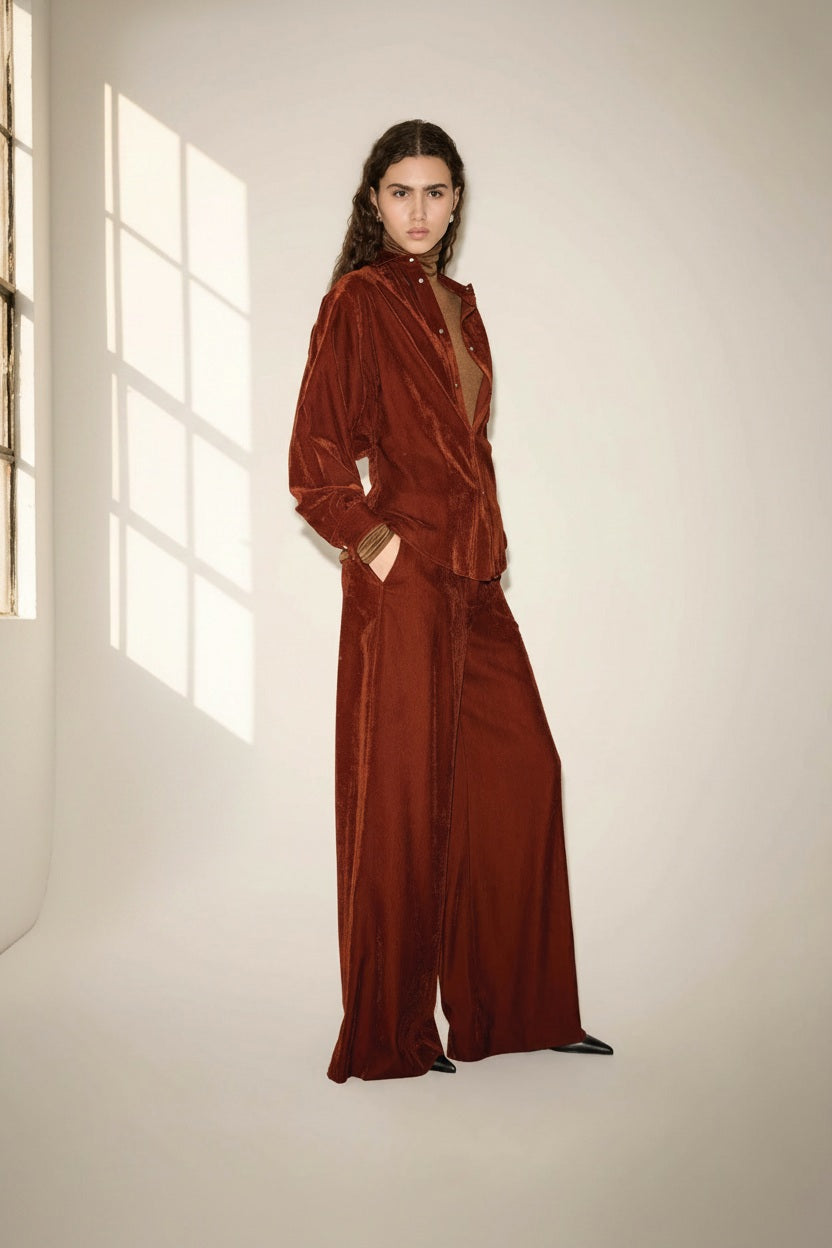A person with long wavy hair stands indoors wearing a loose-fitting, rust-colored velvet jacket paired with the NUDE - WIDE LEG TROUSER IN TOBACCO by NUDE. Crafted from soft viscose baby cord, the high-rise trousers flatter their silhouette as they rest their hands in the jacket pockets. Soft natural light streams through a window, casting gentle shadows on the wall.