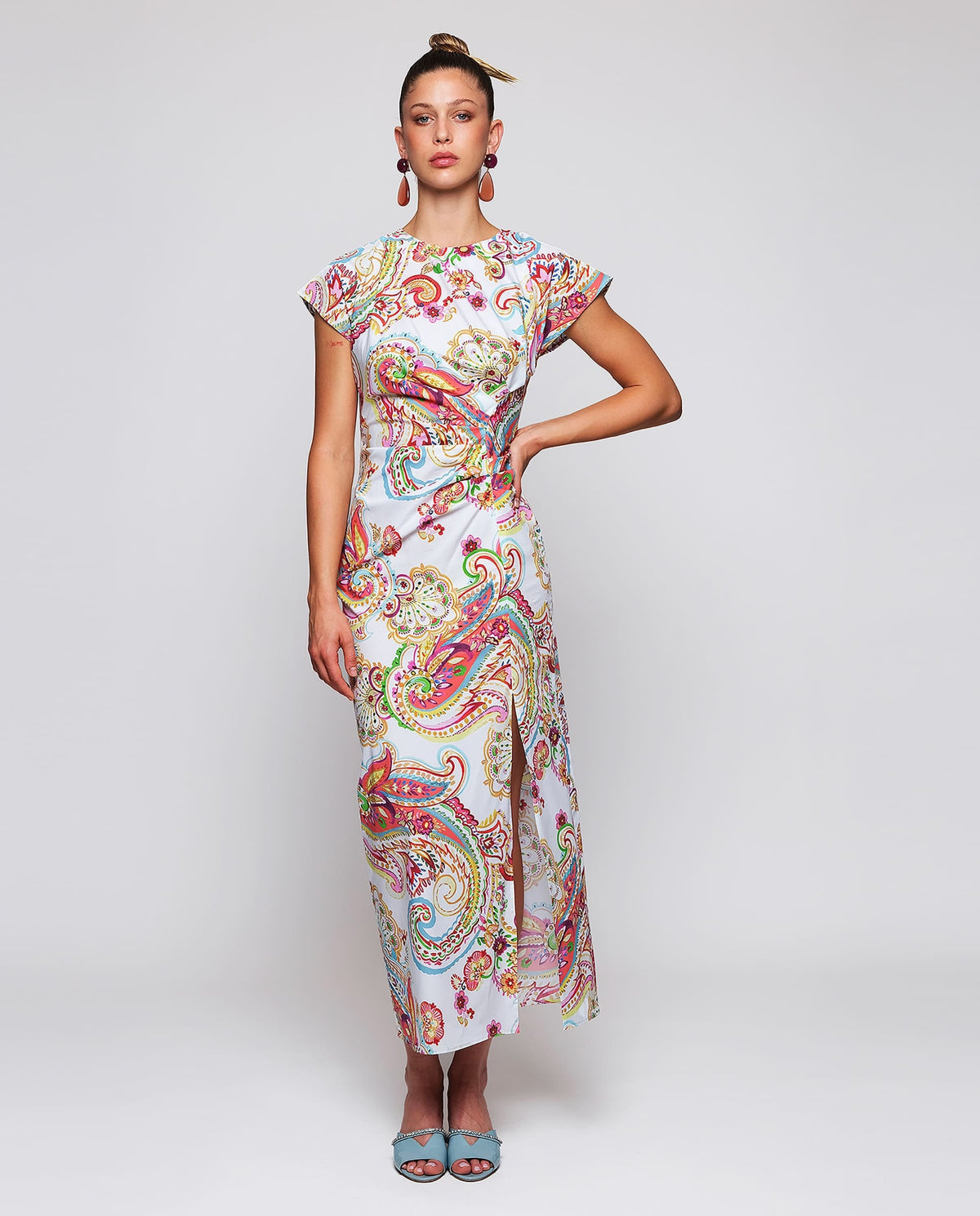 A woman stands against a white background wearing the MIRTO 1956 - PAISLEY PRINTED MIDI DRESS with short sleeves and light blue open-toe heels, her hair in a high bun.