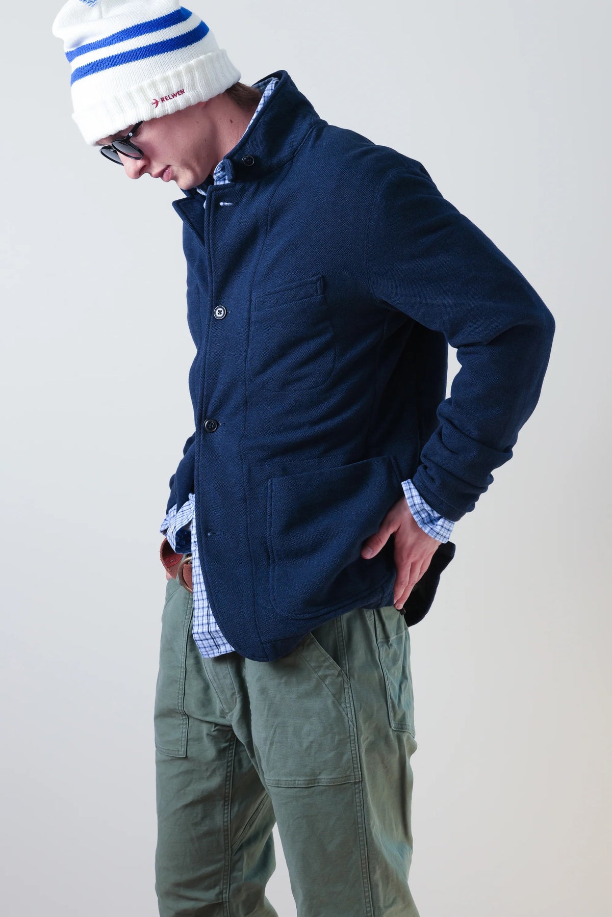 A person wearing the RELWEN - PIQUE TRAP BLAZER IN NAVY HEATHER over a checked shirt and green cargo pants stands with hands in pockets against a plain background, showcasing the blazer’s relaxed fit.