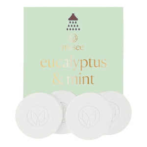 A pastel green packet labeled “MUSEE - EUCALYPTUS & MINT SHOWER STEAMERS” from MUSEE, featuring a shower icon, is displayed behind four round white shower steamers embossed with a leaf design—offering a spa-like experience with these natural bath products.