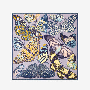 A woman with straight brown hair pulled back wears a dark top and drapes the INOUI EDITIONS PAPILLON (BUTTERFLIES) Square Scarf in Grey by INOUI, featuring abstract and butterfly patterns, around her neck against a white background.