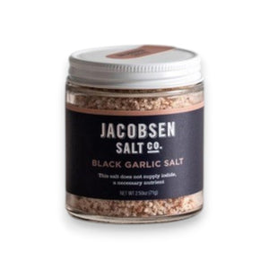 A jar of JACOBSEN SALT CO. INFUSED BLACK GARLIC SALT, featuring a black label and white lid, rests on a light beige surface against a plain light gray background.