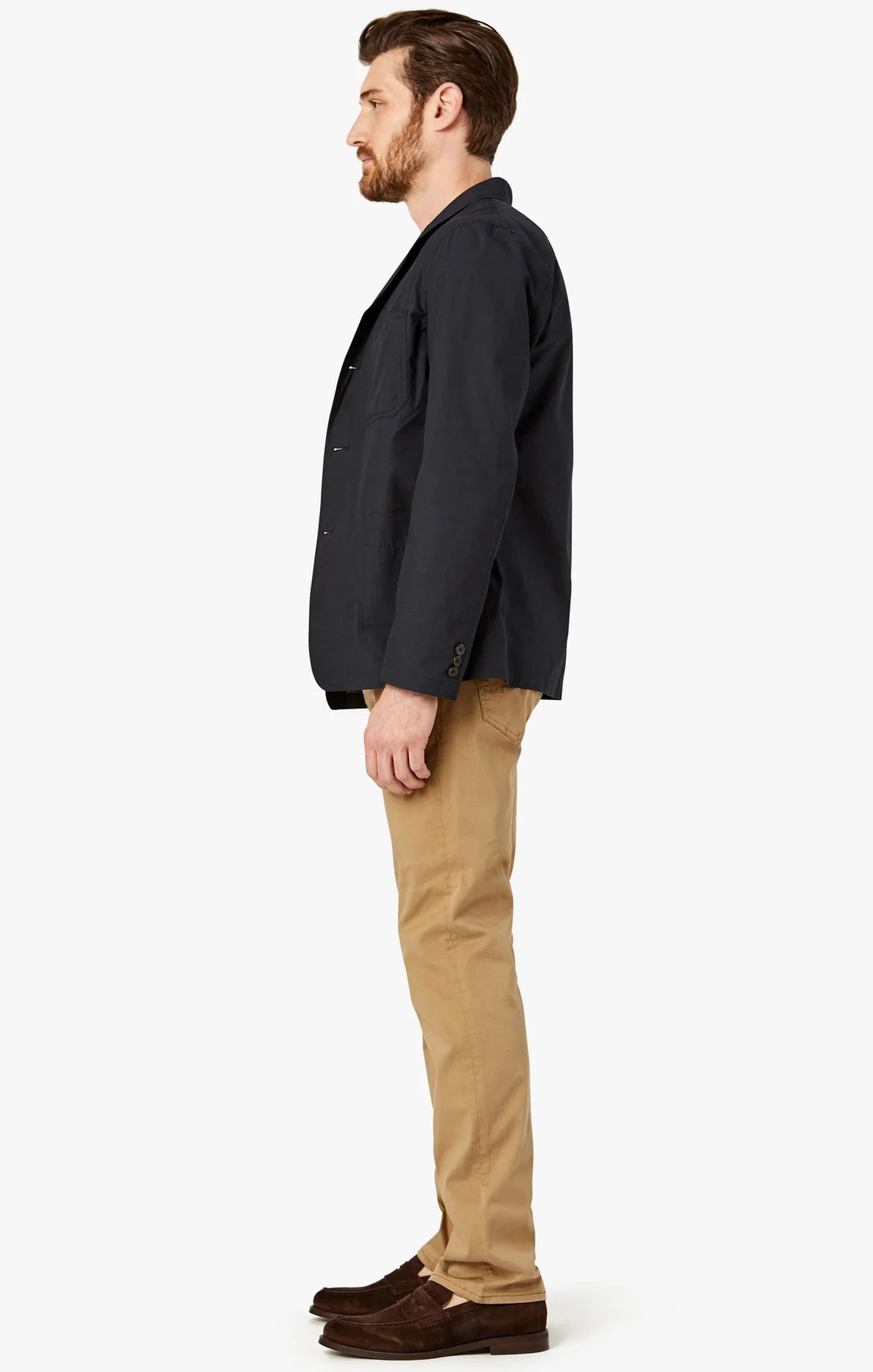 A man stands against a white background wearing 34 HERITAGE - COURAGE STRAIGHT LEG PANT IN KHAKI TWILL, paired with a navy blazer, a white and navy striped shirt, and brown shoes, looking to his left with one hand in his pocket.