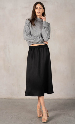 A woman in a sleeveless black dress with a wide belt stands against a plain backdrop, holding a large black handbag. She wears black shoes and layers the ROSE AND CROWN - SATIN RIBBED SKIRT by ROSE & CROWN under her dress.