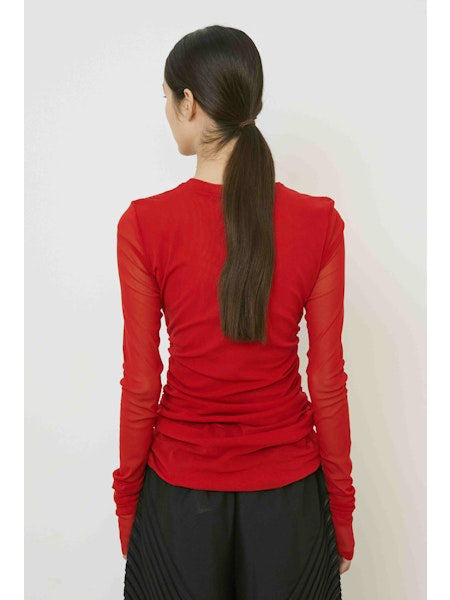 A woman with long dark hair in a low ponytail stands in profile against a plain light background, wearing the MEIMEIJ MESH LAYERING TOP by MEIMEIJ in red and a black skirt for a minimalist Italian fashion look.