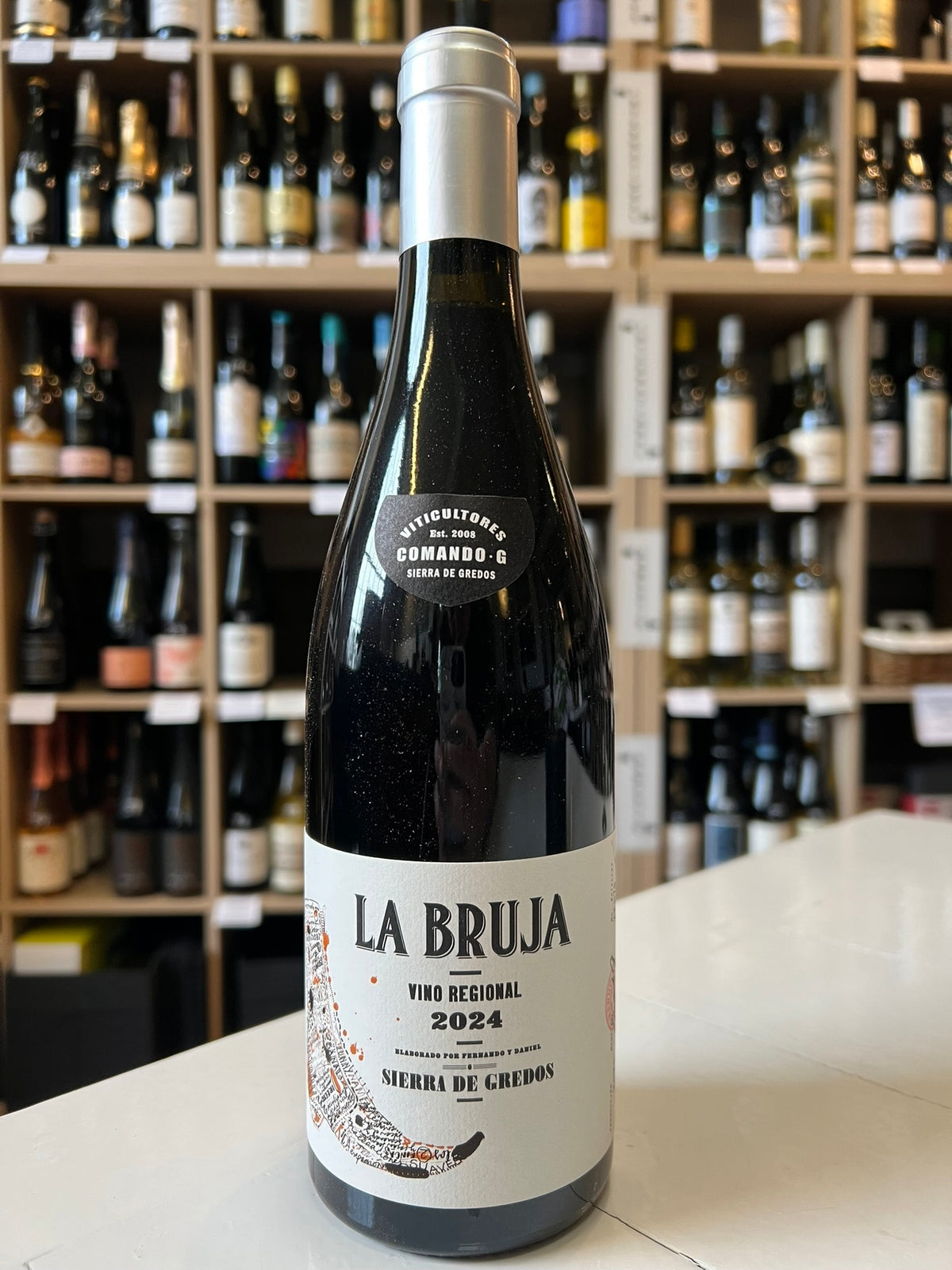 A bottle of COMANDO G 'La Bruja' Grenache 2024 by Wine: Freedom Beverage stands on a counter before shelves of other wines. The bottle features a white label with black and red text.