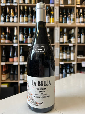 A bottle of COMANDO G 'La Bruja' Grenache 2024 by Wine: Freedom Beverage stands on a counter before shelves of other wines. The bottle features a white label with black and red text.