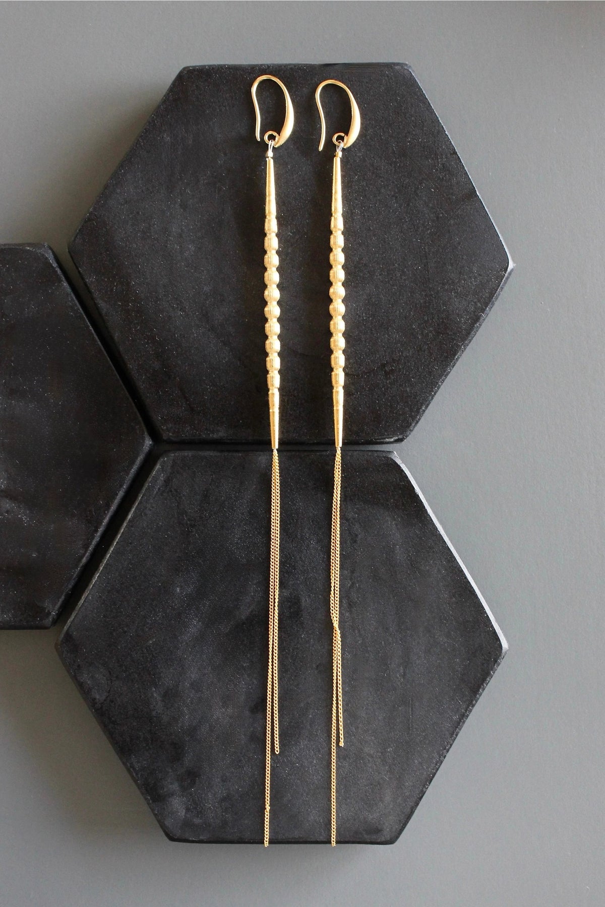 DAVID AUBREY - GOLD PLATED SHOULDER DUSTER EARRINGS feature small round beads and delicate gold-plated chain ends, elegantly displayed on black hexagonal tiles set against a gray background.