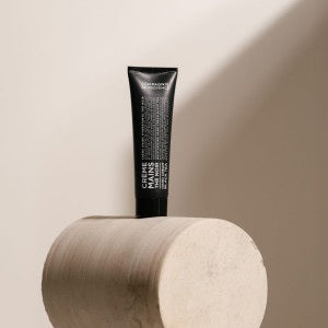 A black tube of COMPAGNIE DE PROVENCE - TRAVEL HAND CREAM BLACK TEA 1OZ from CIE LUXE BRANDS stands upright on a light, cylindrical stone pedestal against a neutral beige background with soft lighting.