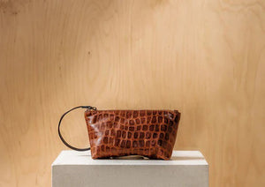 The DANIELLA LEHAVI - LORRIE CLUTCH, a small brown leather wristlet with a crocodile pattern, is displayed on a white pedestal against a light wooden background, highlighting its sophisticated Italian craftsmanship.