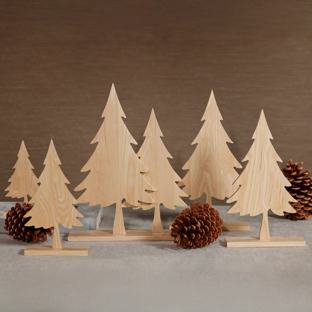 Six ASH WOOD SILHOUETTE TWO TREES ON BASE ornaments, arranged with pinecones on a tabletop.