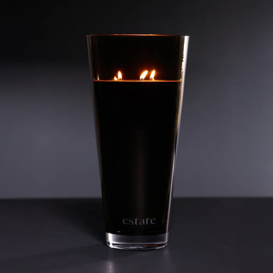 A collection of ZODAX luxury scented candles, including the 57 OZ THISTLE ESTATE CANDLE in black and smoky brown, is displayed on a white textured surface against a plain light background.