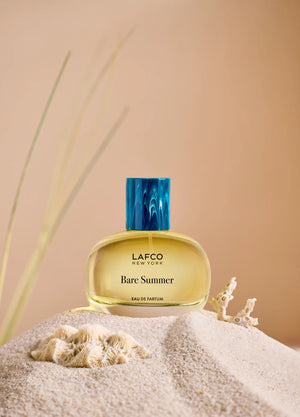 The LAFCO - FINE FRAGRANCE, BARE SUMMER 50ML perfume bottle, featuring a blue cap and displayed alongside delicate grasses, showcases its fresh and uplifting scent inspired by the sea and sand dunes. The text highlights opening, heart, and base fragrance notes that evoke the essence of summer. This vegan scent from LAFCO offers a clean, natural allure perfect for warm days.