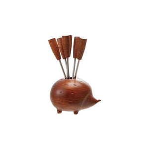 The CREATIVE COOP WOOD HEDGEHOG HOLDER WITH 6 APPETIZER PICKS sits on a tray with a folded napkin, atop two stacked books on a rustic wooden table against a neutral background.
