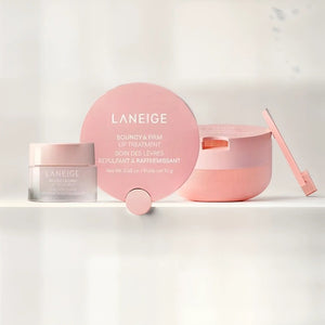 A pink LANEIGE - BOUNCY & FIRM LIP MASK (12g) by KOSMIC KBEAUTY SKINCARE sits open with its lid off, displaying the product inside. Beside the round container lies a small applicator. The label reads “Bouncy & Firm Lip Mask.” Designed to plump and moisturize dry lips overnight, this lip mask leaves your lips soft and healthy-looking.