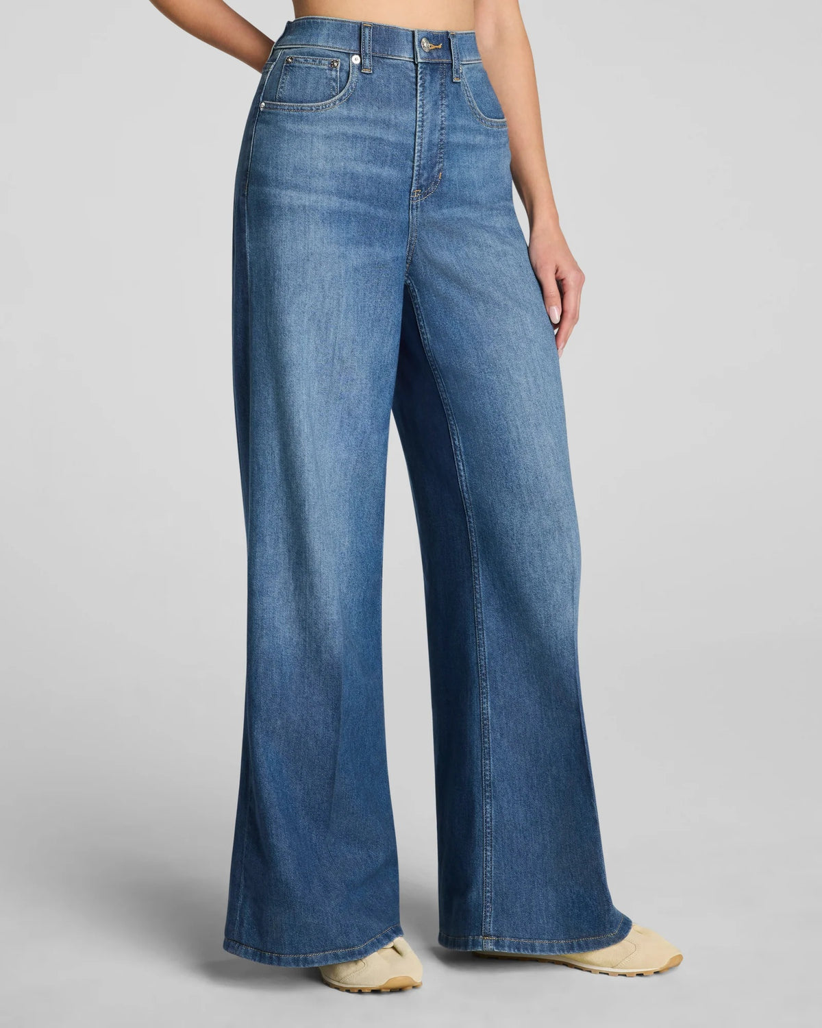 A woman with long brown hair stands against a plain background in a relaxed pose, one hand in her pocket. She is wearing a sleeveless white top, light-colored shoes, and the SPANX SPANXSUPERSMOOTH Authentic Lightweight Super Wide Leg Jean in Oceanic by SPANX, featuring a flattering tummy-smoothing fit.