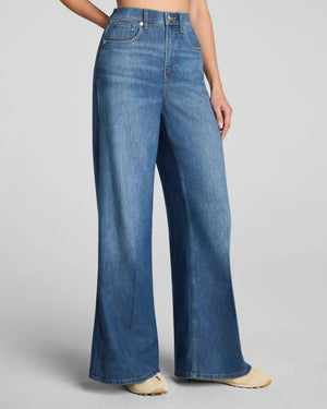 A woman with long brown hair stands against a plain background in a relaxed pose, one hand in her pocket. She is wearing a sleeveless white top, light-colored shoes, and the SPANX SPANXSUPERSMOOTH Authentic Lightweight Super Wide Leg Jean in Oceanic by SPANX, featuring a flattering tummy-smoothing fit.