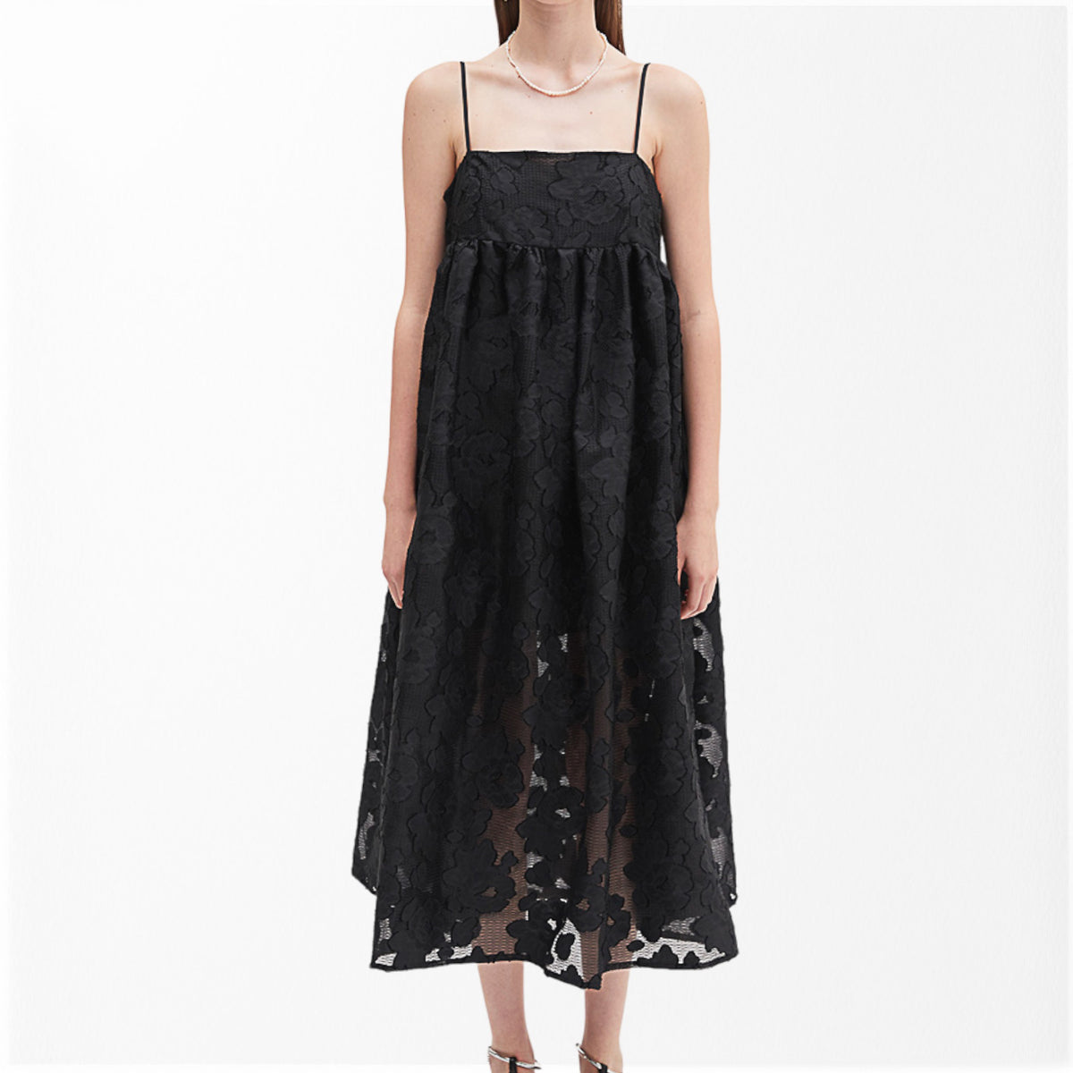 A woman wears the MEIMEIJ Lace Empire Waist Dress, a sleeveless black floral lace midi with spaghetti straps and a sheer overlay. The flowing fit makes it perfect for a garden party. She stands against a plain white background.