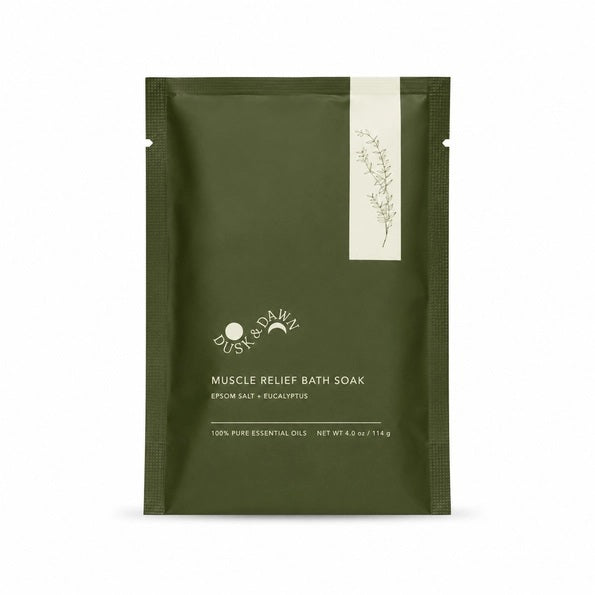 A green pouch of GIFTEN MARKET MUSCLE RELIEF BATH SOAK - EUCALYPTUS OIL rests on the edge of a bathtub lined with white tile, as water pours from a gold faucet in the foreground. Formulated with soothing eucalyptus oil and Epsom salt, this bath soak is an ideal choice for unwinding after a long day or adding to a thoughtful gift basket.