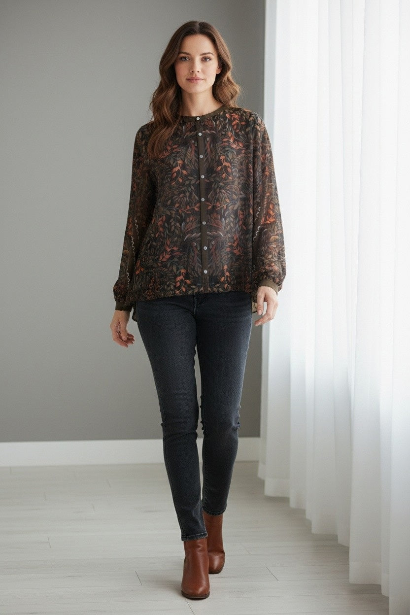 A woman with shoulder-length brown hair and a neutral expression stands indoors wearing the FRANCO FERRARI GERRY Silk Top, a sheer, long-sleeved piece featuring a vibrant botanical print, paired with a matching skirt.