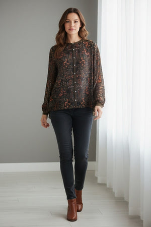 A woman with shoulder-length brown hair and a neutral expression stands indoors wearing the FRANCO FERRARI GERRY Silk Top, a sheer, long-sleeved piece featuring a vibrant botanical print, paired with a matching skirt.