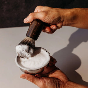 Two round, white BAUDELAIRE - M.B. SHAVING SOAP bars by BAUDELAIRE are displayed. The front bar features an embossed profile of a bearded man, set against a plain white background that evokes the clean, minimalist feel and hints at the smooth razor glide and nourishing shea butter found in each bar.