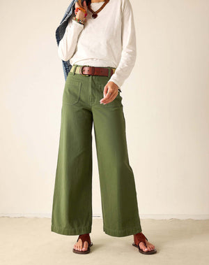 A woman with long brown hair and a relaxed expression stands in a neutral room wearing a denim jacket, white shirt, brown belt, sandals, and holding a tan bag. She pairs her outfit with MERSEA’s Londonberry Twill Pant in Olive for a laid-back look.