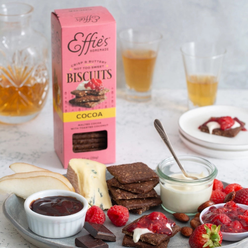 A pink box of EFFIES - COCOA BISCUIT from EFFIES HOMEMADE, labeled “Crisp & Buttery, Not Too Sweet.” The packaging displays chocolate biscuits with raspberries and includes a window to showcase the rich malted cocoa biscuits inside.