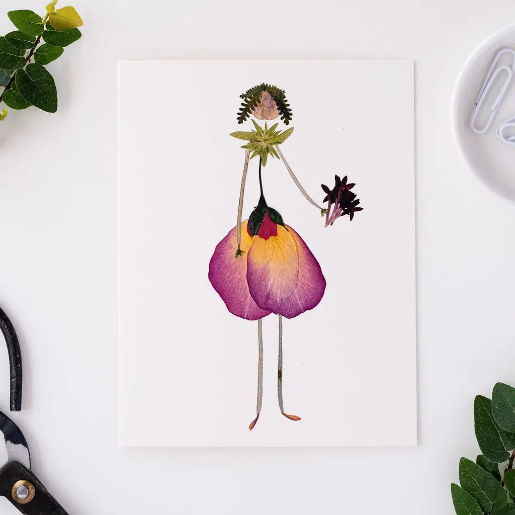 The PETAL PEOPLE PRESS - THINKING OF YOU CARD by PETAL PEOPLE PRESS features a person illustrated with pressed flowers and leaves, petals as a dress, and stems as arms and legs, displayed on a white table with greenery and crafting tools.