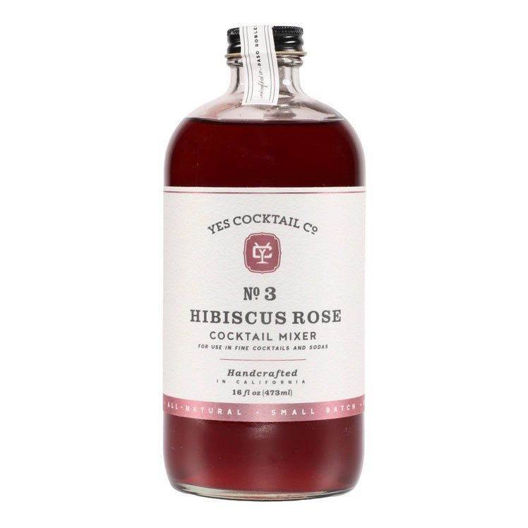 A transparent glass bottle holds a rich, dark red liquid labeled "YES COCKTAIL CO - Hibiscus Rose Cocktail Mixer." Handcrafted in California and available in a 16 fl oz (473ml) size, the white-and-red-toned label indicates its perfect suitability for creating cocktails or an exotic floral spritzer.