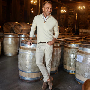A man wearing the COLLARS AND CO Luxury Touch Cotton and Cashmere Quarter Zip Sweater in cream with brown suede trim stands smiling with crossed legs, leaning on a wine barrel in a dimly lit cellar lined with barrels.