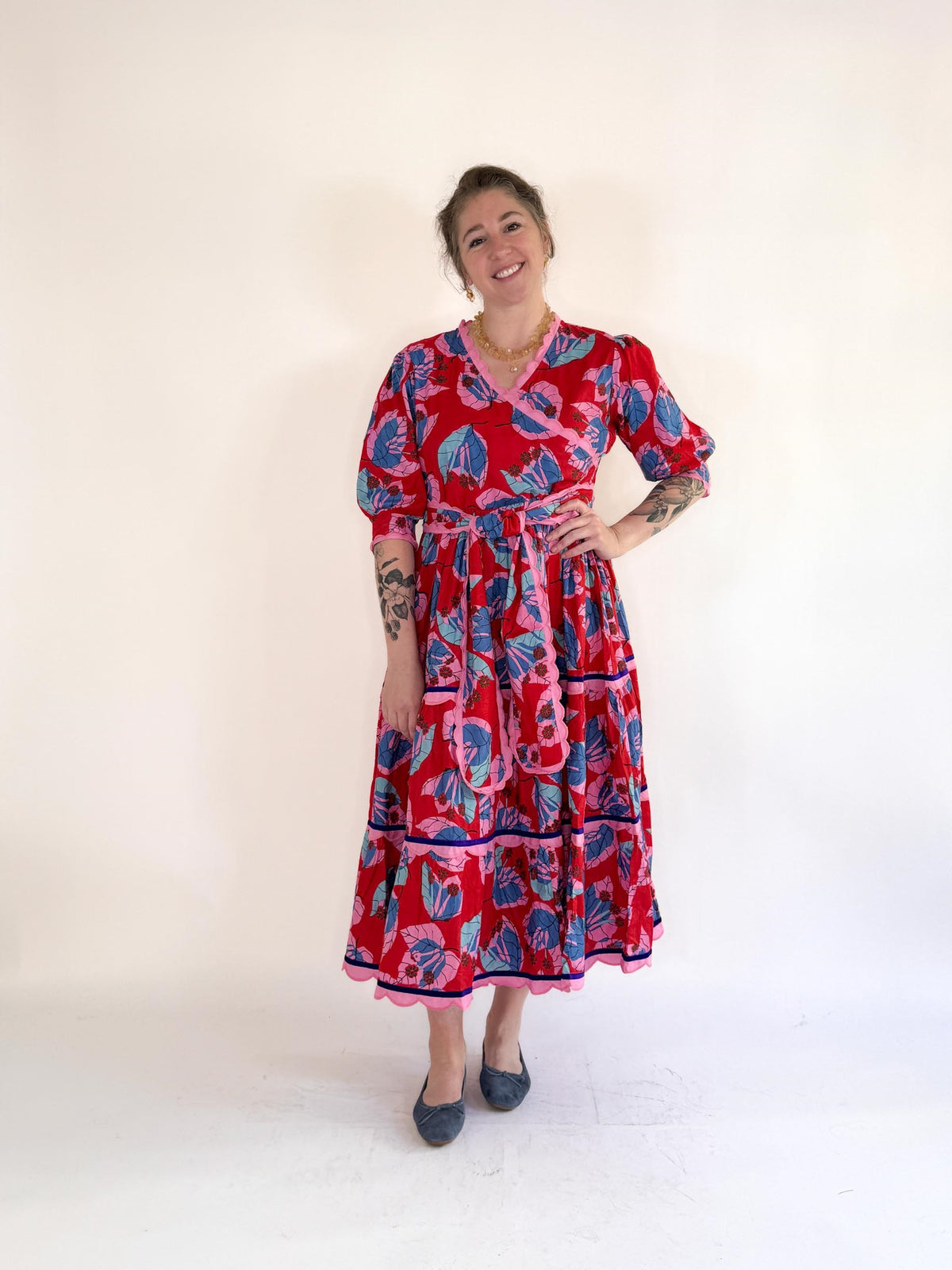 A smiling woman in the FARYLINA - CAMILLE LONG DRESS, a bold blue and pink floral cotton dress by FARYLINA, stands against a white background with one hand on her hip, showing arm tattoos and wearing gray flats.
