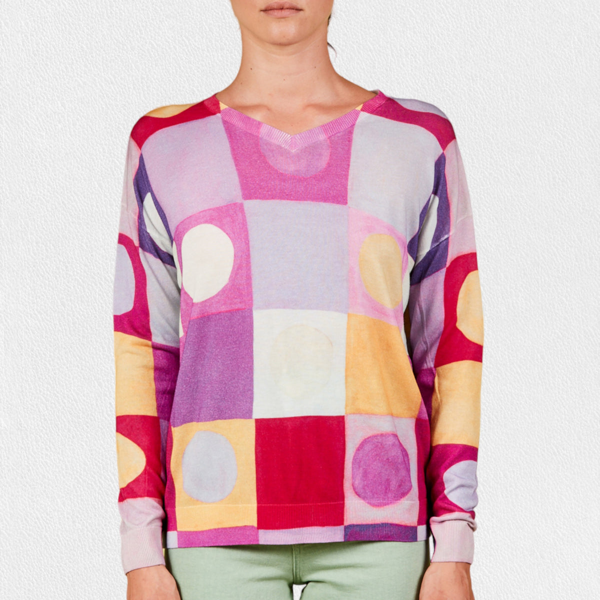 A person wearing the IN BED WITH YOU - BATWING SLEEVE V NECK JUMPER stands against a plain, light-colored background. The colorful geometric pattern featuring squares and circles in shades of pink, purple, red, yellow, and white adds flair to this stylish piece by the Italian brand IN BED WITH YOU/CLAUDIOLAI SRL. The person is visible from the shoulders down to the waist.