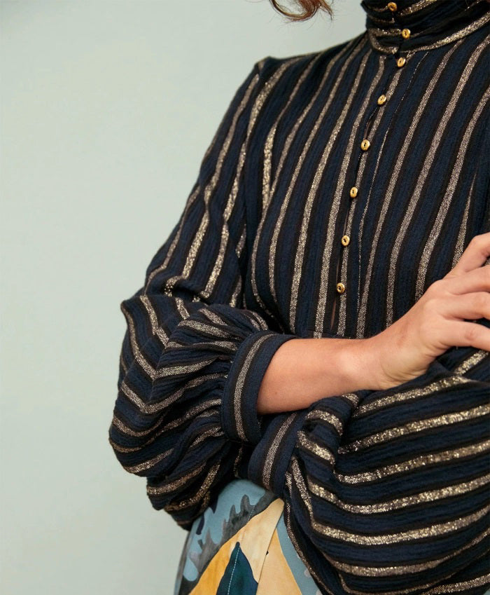 A woman with wavy brown hair, arms crossed, wears the MOMONI Thierry Blouse in lurex stripe gauze and a patterned skirt, standing against a pale background.