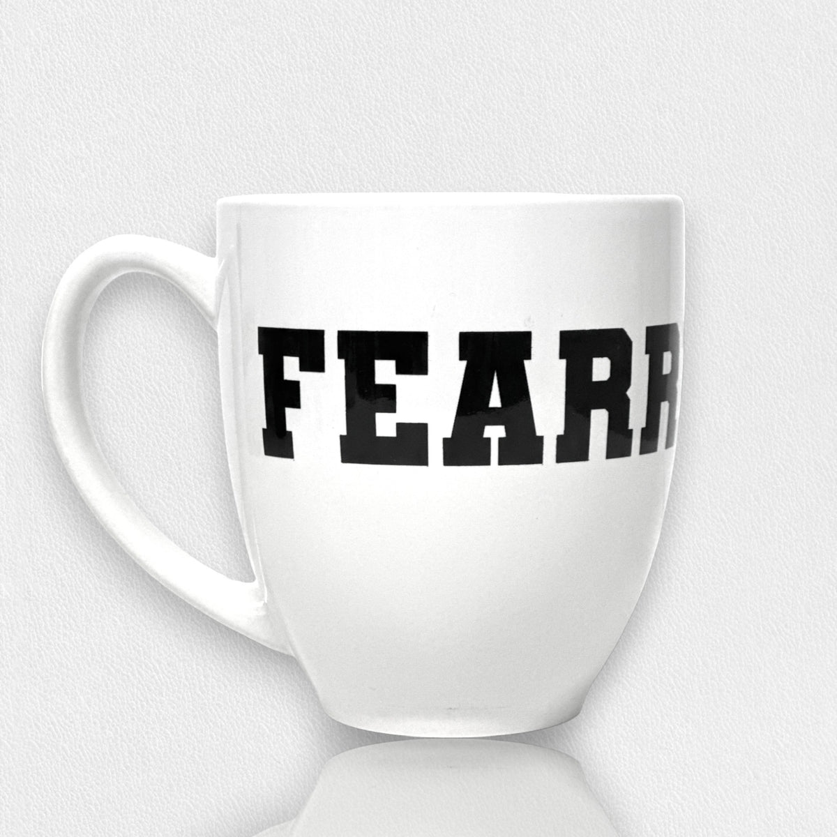 The FEARRINGTON LIFESTYLE COLLECTION - FEARRINGTON MUG by STRAN features the word &quot;FEARLESS&quot; boldly displayed in black, capital letters across its front. Perfect for your morning coffee, this ceramic mug rests on a white surface with a subtle shadow. The clean and minimalistic backdrop highlights its sleek design.