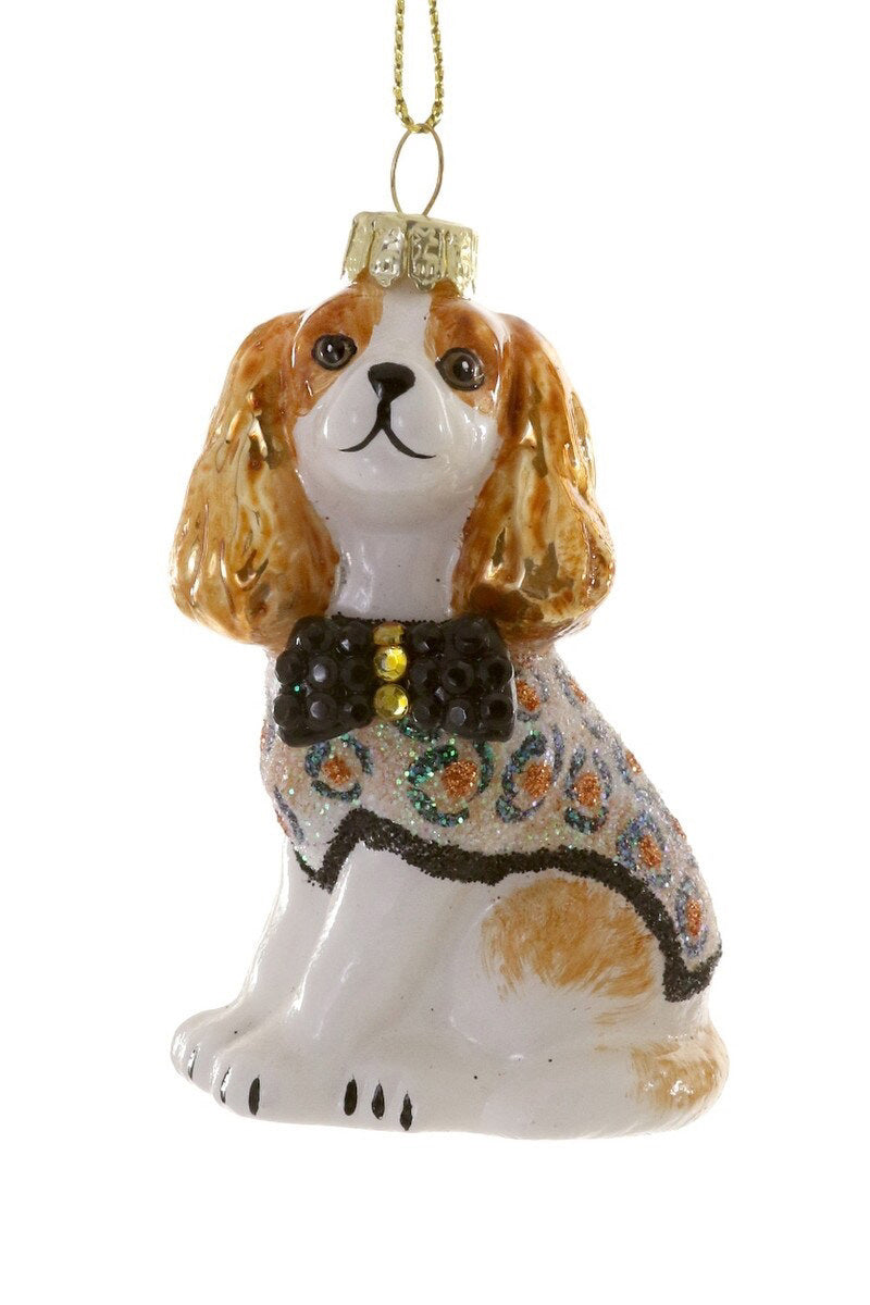 Introducing the SOPHISTICATED SPANIEL ORNAMENT by CODY FOSTER: a charming glass Christmas decoration shaped like a dog wearing a glittery sweater and a black bow tie. Featuring a gold loop for hanging, this festive ornament showcases brown ears and a white body adorned with intricate patterns on its sweater, adding holiday cheer to any height on your tree.