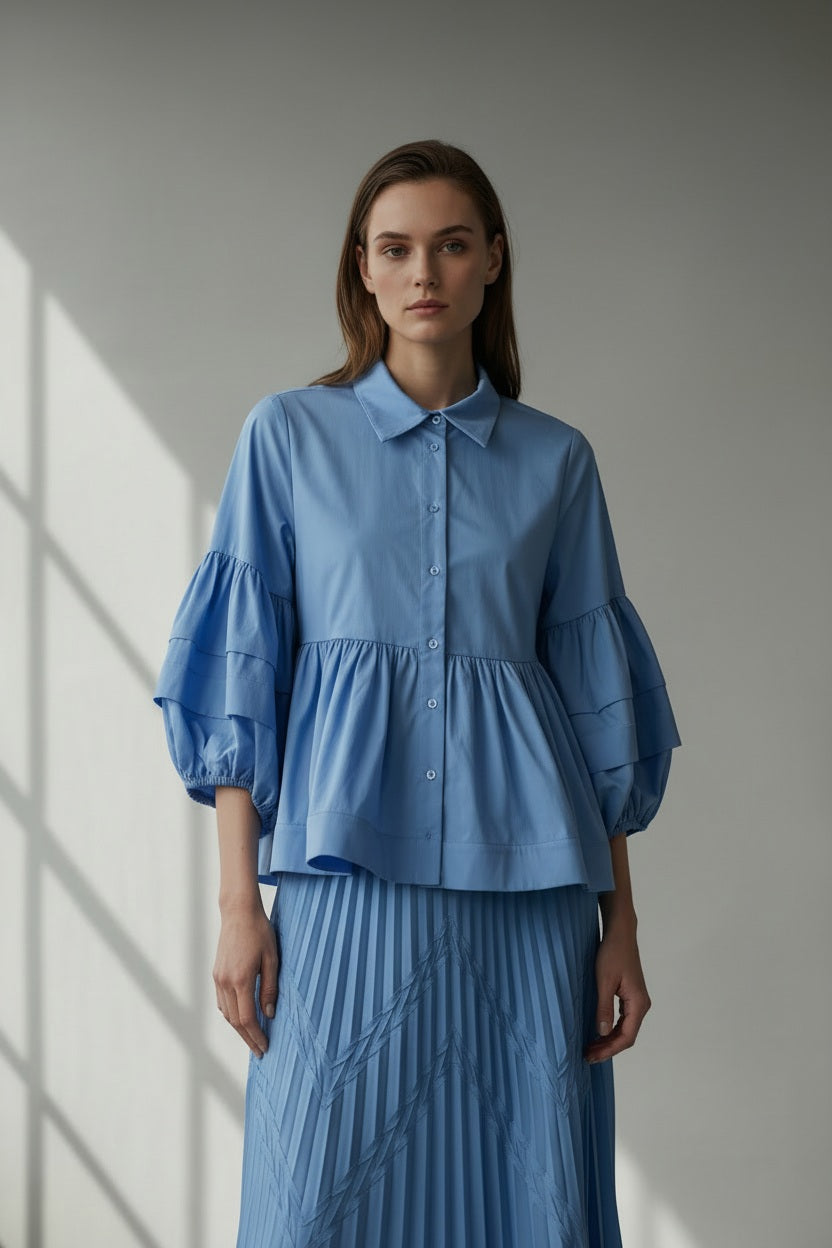 A woman with straight, light brown hair wears the MILVA MI - COTTON BLOUSE IN AZZURO BLUE by MILVA, featuring voluminous sleeves and a peplum hem, paired with bold white earrings against a plain gray background.