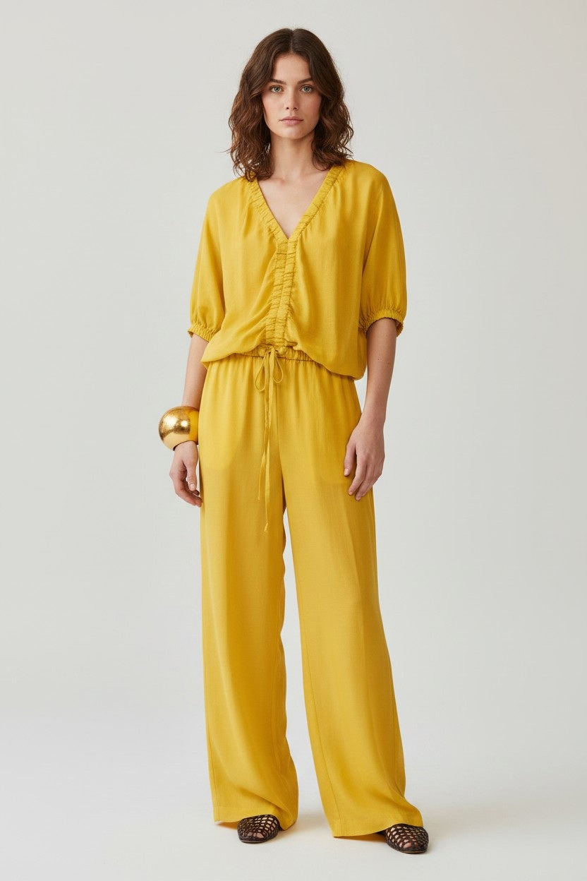 A woman with wavy brown hair wears a loose, bright yellow V-neck ruched blouse and NUDE's WIDE LEG PANT, crafted from a Tencel-cotton blend. She stands against a plain background, accessorized with a chunky gold bangle.