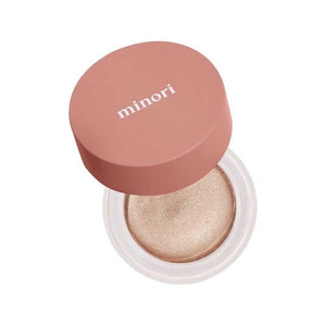 A small round container of MINORI - CREAM HIGHLIGHTER, CHAMPAGNE with a pink lid is shown partly open, revealing the shimmery champagne cream inside for a natural glow.