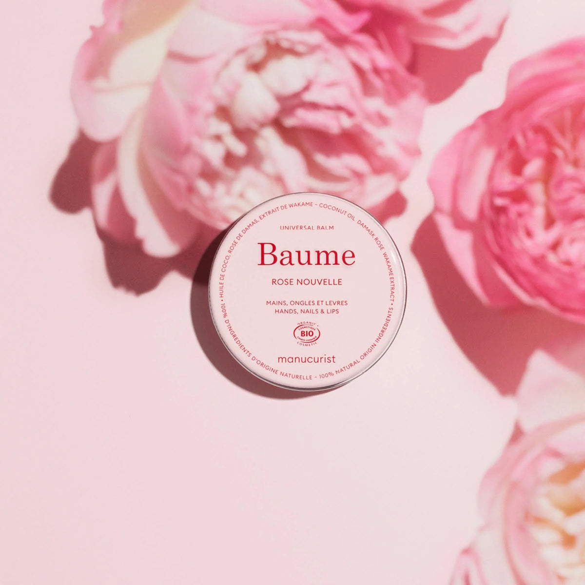 With red nails, a person with medium-dark skin applies MANICURIST - ROSE BALM COSMOS ORGANIC NATURAL NOURISHING BALM by MANUCURIST INC to the back of their left hand against a light pink background. Perfect for lips, nails, and cuticles.