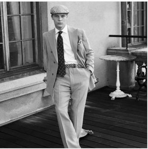A man in a suit and patterned flat cap wears the WIGENS - SIGNATURE WOVEN BELT IN SAND as he leans casually against a window ledge on a wooden patio, with a small round table and decorative chair in the background.